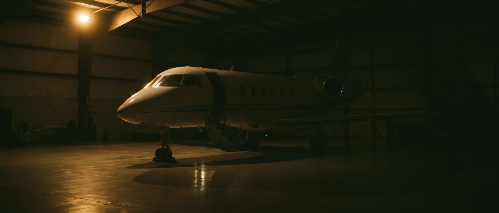Kadr artystyczny: odrzutowiec biznesowy w ciemnym hangarze nocą, jedna ciepła lampa robocza na kadłubie